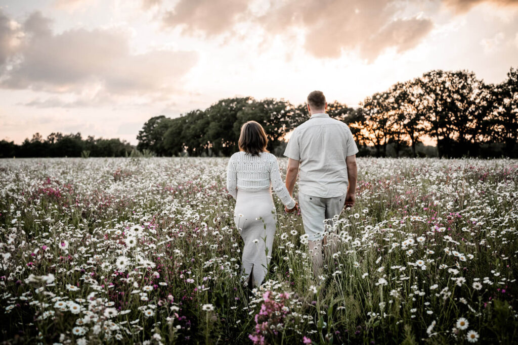 Paar läuft Hand in Hand mit dem Rücken zugewandt im Sonnenuntergang über eine große Blumenwiese, tragen heille, beige Kleidung, warum babybauchshooting machen